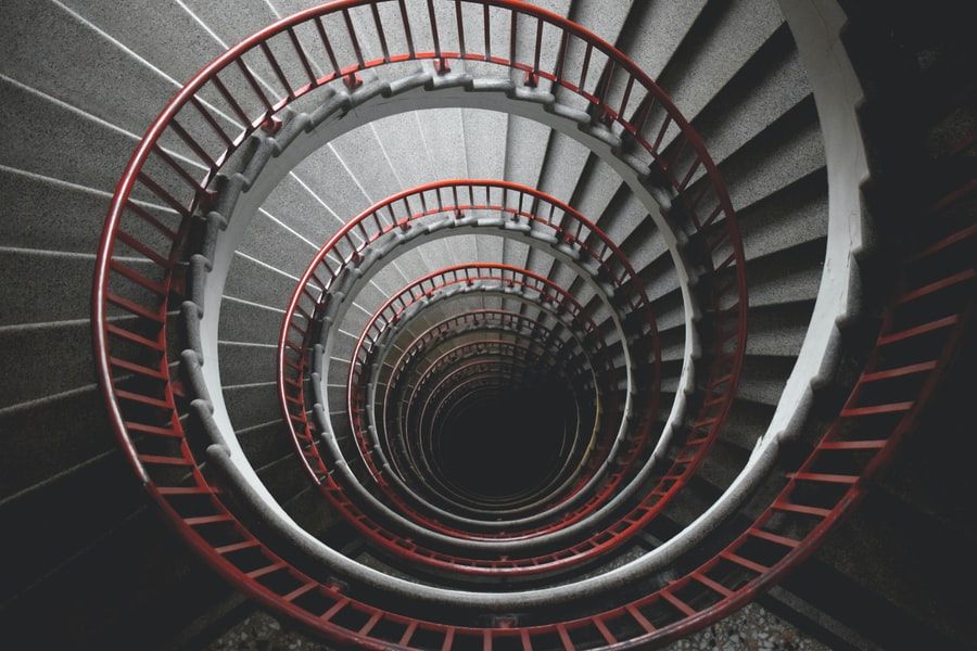 Looking down the middle of a long spiral staircase with a red barrier, descending into darkness, where the bottom is not visible.