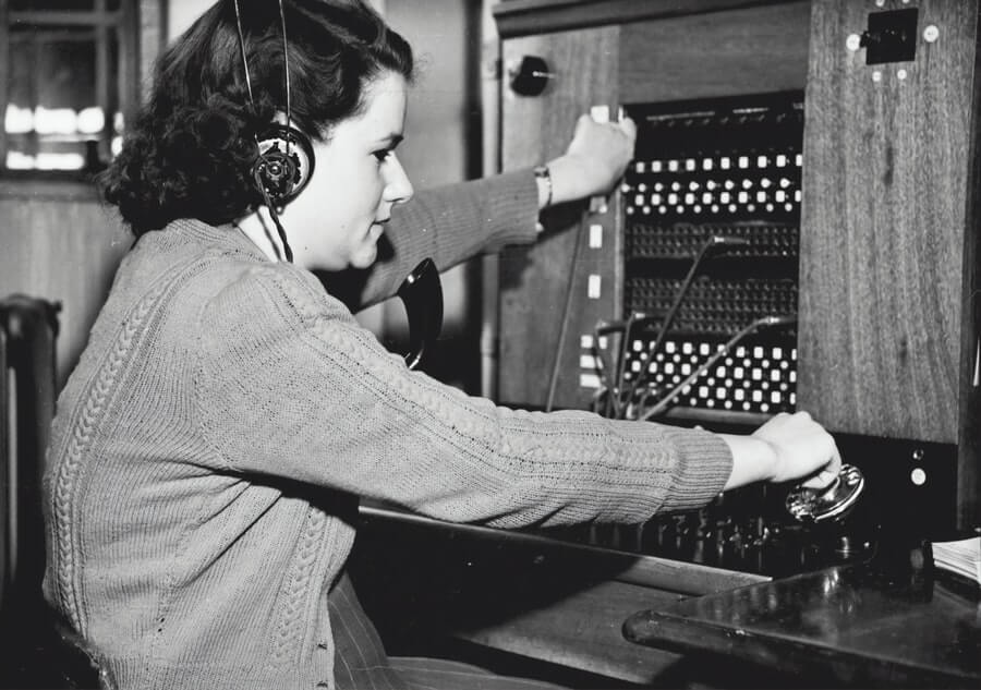 A woman sits at and operates an old phone switchboard. Photo is in black and white.